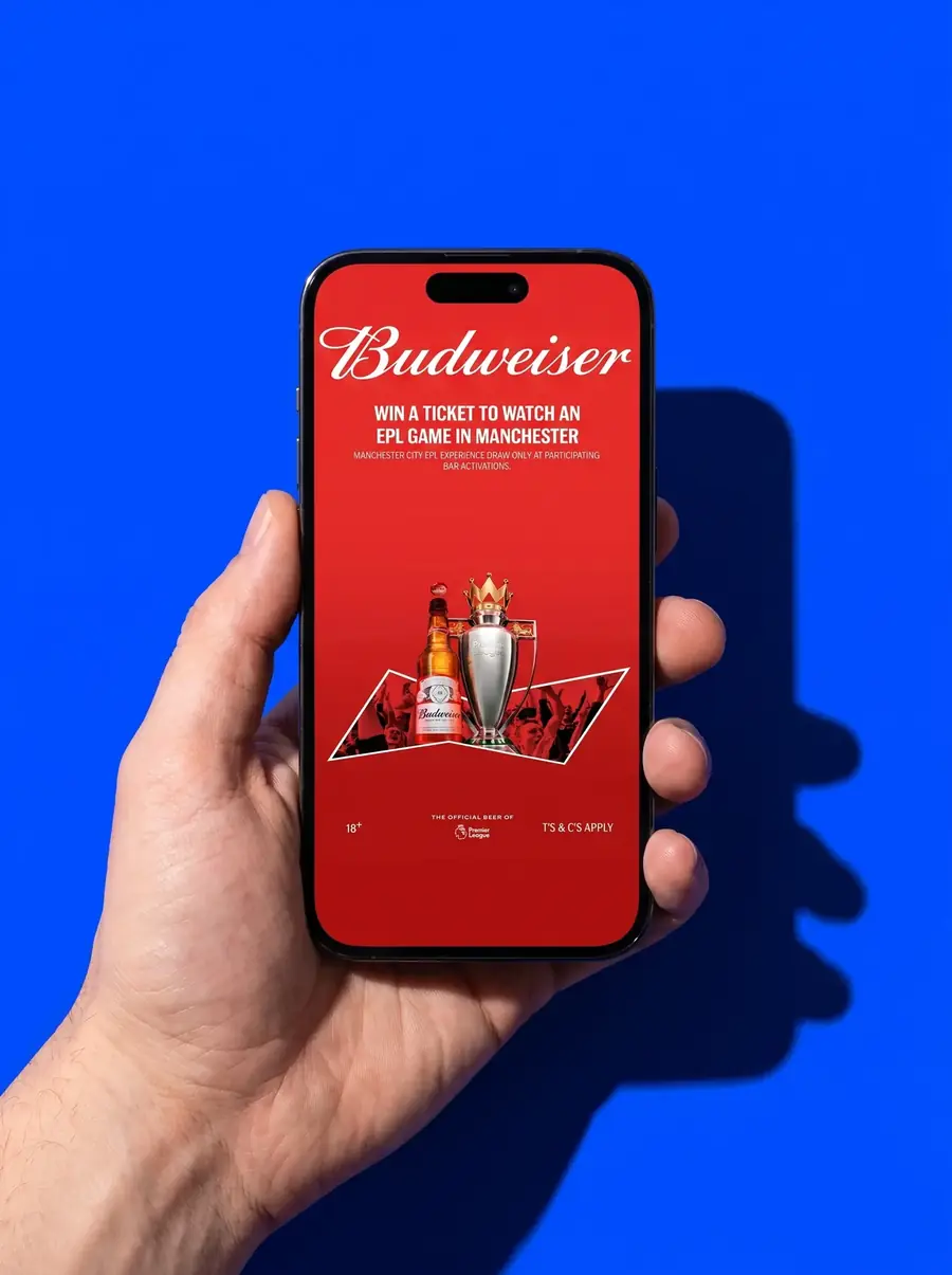 A hand holds a phone on a blue backdrop showing the Budweiser activation: win a ticket to an EPL game