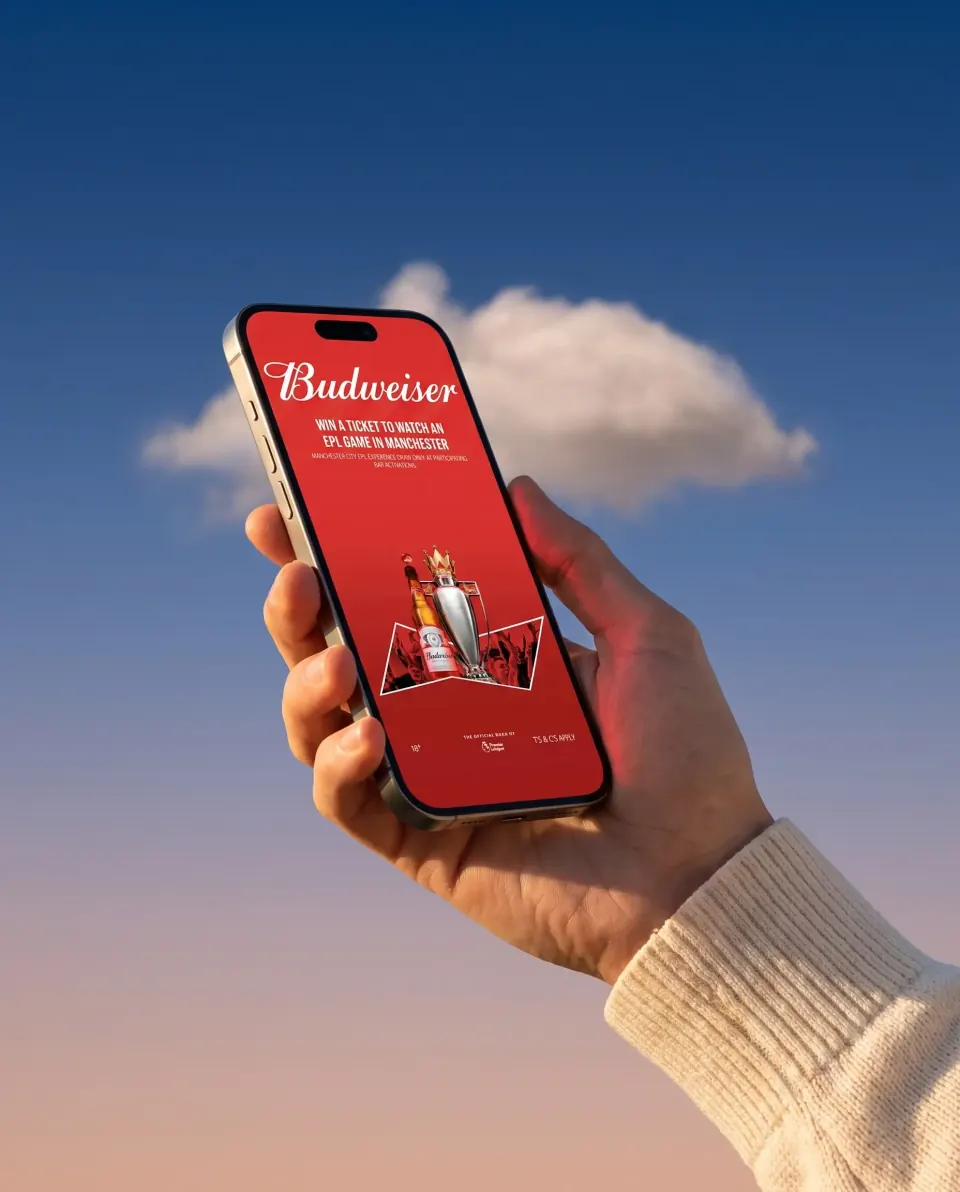 A phone raised against an evening sky showing the Budweiser activation: win a ticket to watch an EPL game in Manchester