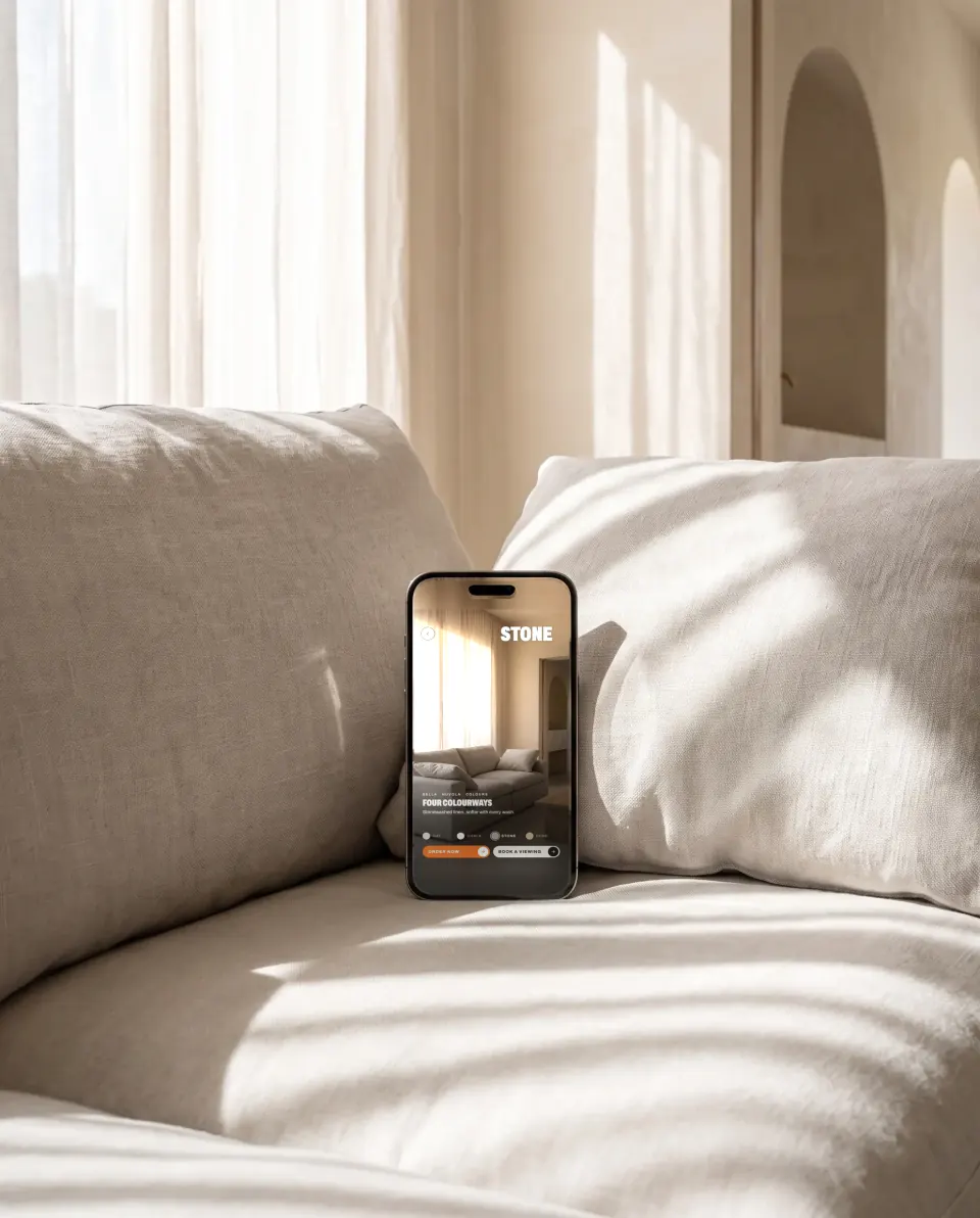A phone between oat linen cushions in morning sun, showing the same sofa in a shoppable video