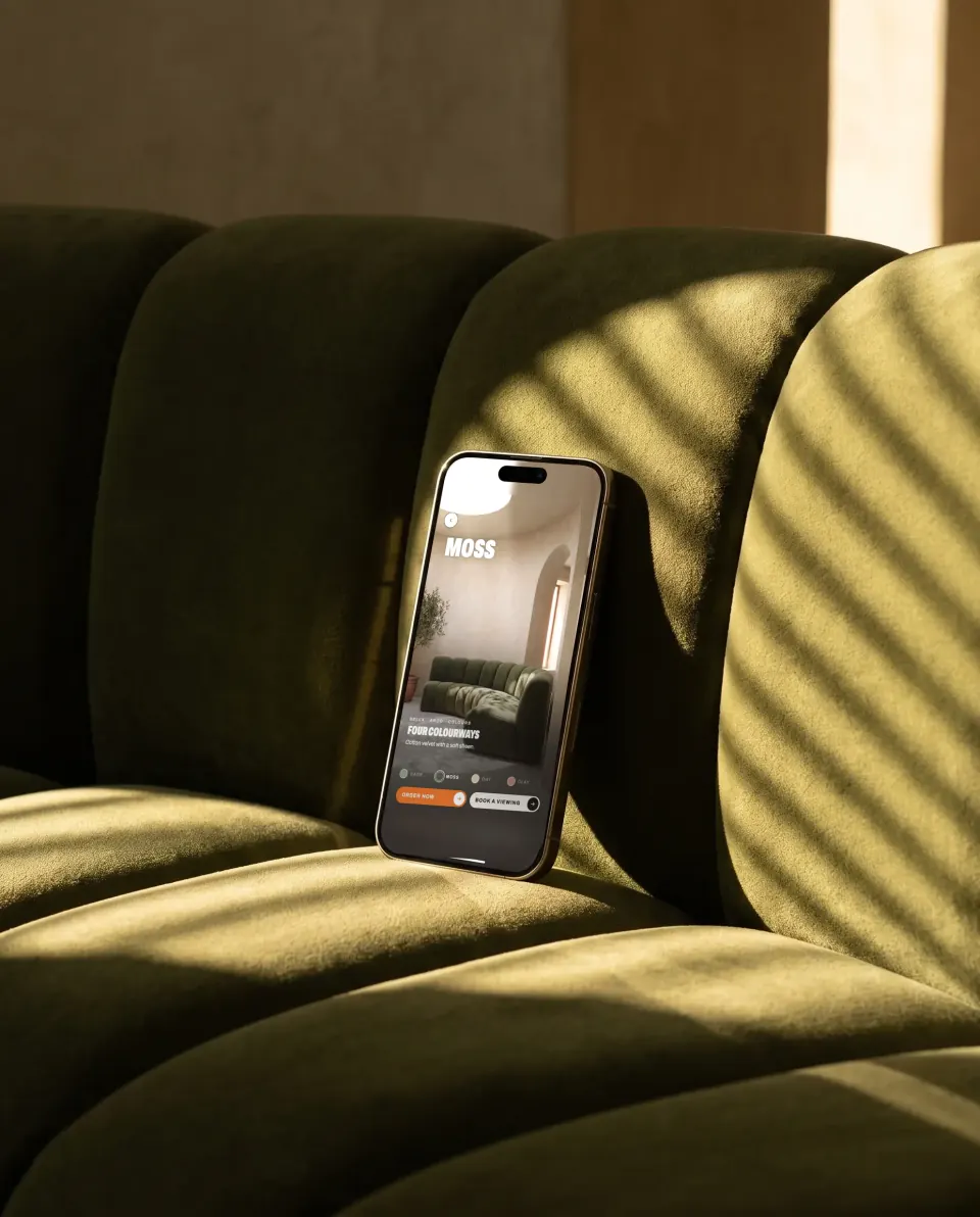 A phone leaning on a moss green velvet sofa in low sun, showing the same sofa in a shoppable video colour picker